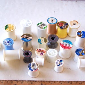 May include: A collection of vintage sewing thread spools in various colors and sizes. The spools are made of plastic and wood, with labels featuring different brand names and designs. Some spools are white, yellow, brown, and blue.