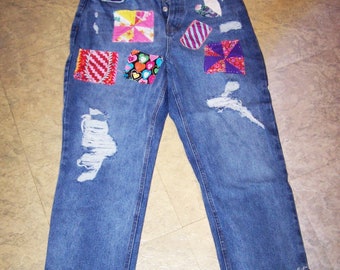 May include: Blue denim jeans with frayed hems and various colourful fabric patches sewn on the front. The patches are in different shapes and sizes, including squares, triangles, and rectangles.