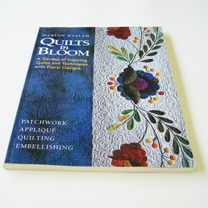 Op de afbeelding: Boekomslag met een blauw en wit bloemenmotief. De titel is "Quilts in Bloom" door Marion Haslam. De ondertitel is "Een tuin met inspirerende quilts en technieken met bloemenmotieven". Het boek bevat ook instructies voor patchwork, appliqué, quilten en versieren.