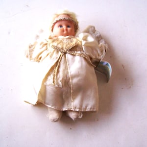 Pode incluir: Boneca de anjo vintage com um vestido de cetim creme e asas a condizer. A boneca tem um rosto de plástico com olhos azuis e uma auréola loira. O vestido é acentuado com acabamentos dourados e detalhes de renda, criando uma estética clássica e nostálgica.