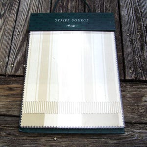 Könnte beinhalten: Ein Stoffmusterbuch mit vertikalen Streifenmustern in Creme- und Beigetönen. Der obere Teil des Buches ist dunkelgrün mit dem Text "STRIPE SOURCE" und einer Unterschrift. Das Buch liegt auf einer verwitterten Holzoberfläche.