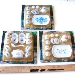 Peut inclure: Trois services à thé miniatures, chacun emballé sur un support en carton. Les ensembles comprennent de petites tasses, soucoupes, théières et plats de service. La vaisselle est blanche avec des motifs floraux bleus ou roses.