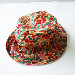 May include: A colorful bucket hat with a repeating pattern of abstract shapes in red, teal, brown, and yellow. The hat has a wide brim and is made of fabric. The hat is a fashion accessory.