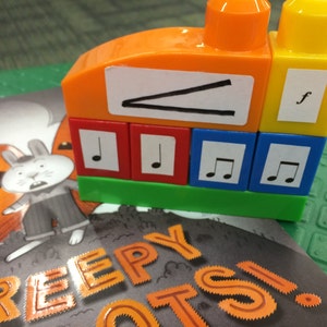 May include: A colorful toy building block set with musical notes and a letter 'f' on the blocks. The blocks are stacked on top of each other and are arranged in a row. The blocks are orange, yellow, red, green, and blue. The blocks are on top of a book with the words 'Creepy Clowns' on the cover.