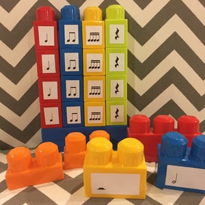 May include: Colorful building blocks with musical notes printed on them. The blocks are red, blue, yellow, and orange. The musical notes include quarter notes, eighth notes, and whole notes. The blocks are arranged in a tower and scattered around.