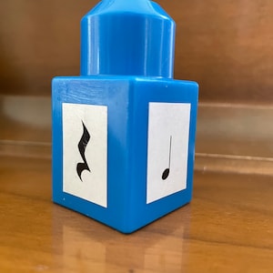 May include: A blue plastic cube with a white square on each side. Each square has a black musical note symbol. One side has a treble clef and the other side has a whole note.