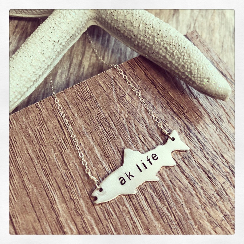 May include: A silver necklace with a fish-shaped pendant that says "ak life". The necklace is on a wooden surface with a starfish in the background.