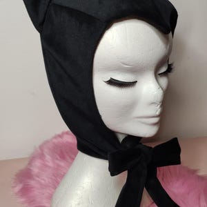 May include: Black velvet cat ear hat with a long black ribbon tied in a bow. The hat is on a mannequin head with a pink fuzzy background.
