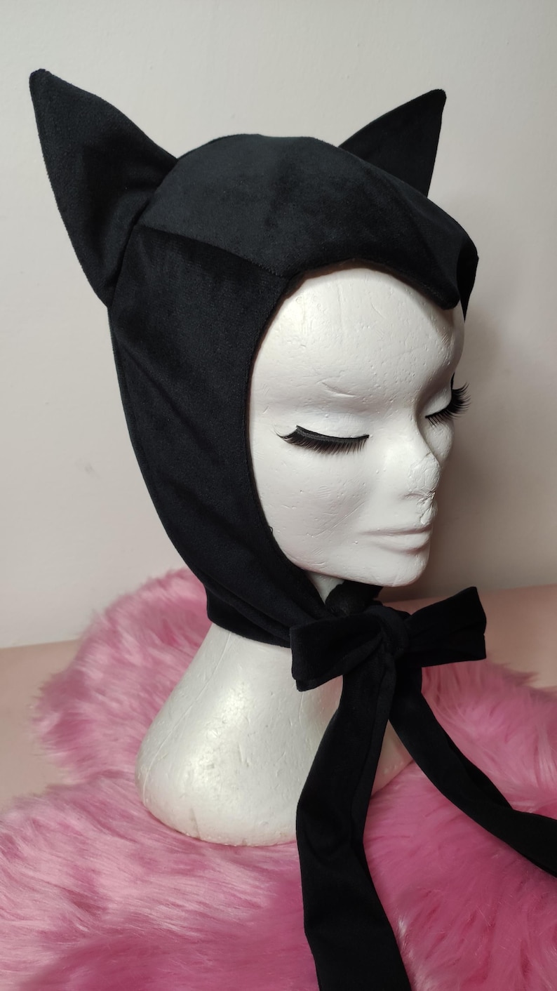 May include: Black velvet cat ear hat with a long black ribbon tied in a bow. The hat is on a mannequin head with a pink fuzzy background.