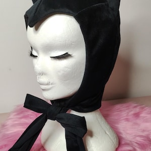 May include: Black velvet cat hood with ears and a black ribbon tie. The hood is on a white mannequin head and is displayed against a pink fuzzy background.