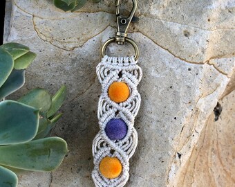 May include: A white macrame keychain with a gold metal clasp and three pom-poms. Two of the pom-poms are orange and one is purple.