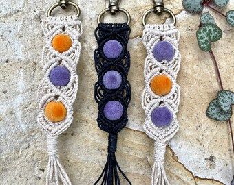 May include: Three macrame keychains with different colored tassels. The keychains are made with white, black, and natural colored string. Each keychain has three purple velvet circles and one orange velvet circle.