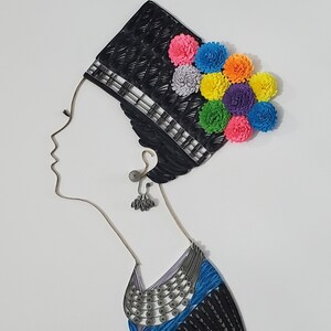 Op de afbeelding: Een kunstwerk van papierquilling dat het profiel van een vrouw toont die een zwarte hoed draagt met een kleurrijk bloemenontwerp en een blauwe en zilveren ketting.