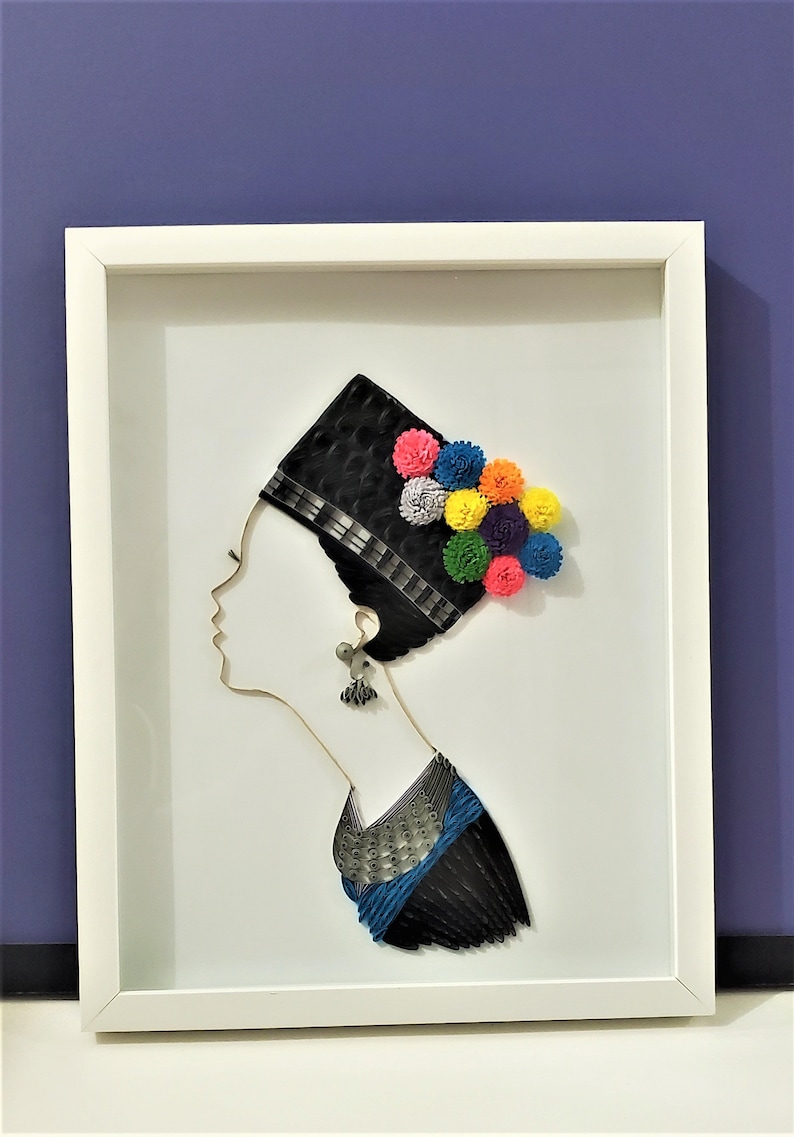 Op de afbeelding: Een wit ingelijst papieren kunstwerk dat het profiel van een vrouw met een zwarte hoed en kleurrijke papieren bloemen toont. De vrouw draagt een blauw en zwart gepatroneerd kledingstuk.