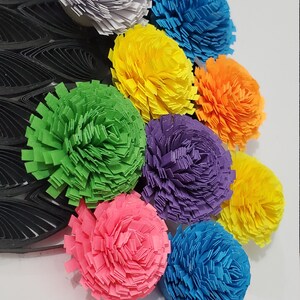 Op de afbeelding: Een verzameling van negen kleurrijke papieren bloemen in verschillende tinten roze, blauw, groen, geel, oranje en paars. De bloemen zijn in een rij gerangschikt, met de grootste bloem bovenaan en de kleinste bloem onderaan.