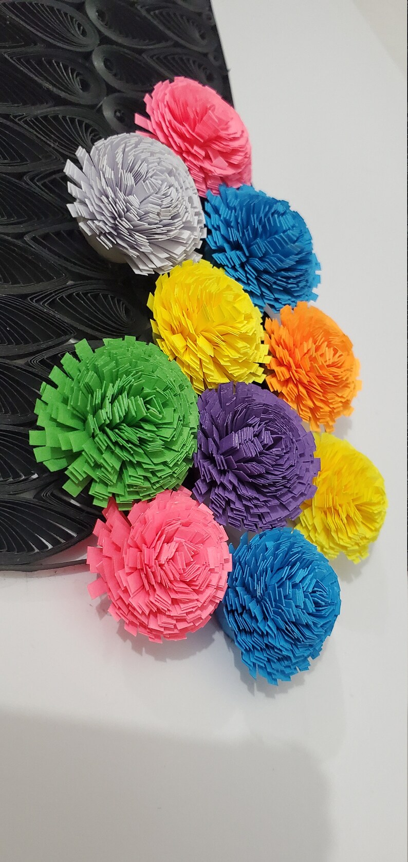 Op de afbeelding: Een verzameling van negen kleurrijke papieren bloemen in verschillende tinten roze, blauw, groen, geel, oranje en paars. De bloemen zijn in een rij gerangschikt, met de grootste bloem bovenaan en de kleinste bloem onderaan.