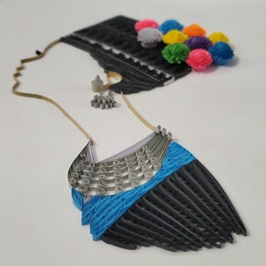 Op de afbeelding: Een kunstwerk van papierquilling dat de silhouet van een vrouw voorstelt met een kleurrijk pompon-haaraccessoire en een blauwe en grijze papierquilling-ketting.