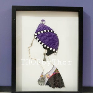 May include: A framed paper art piece depicting a woman's profile wearing a purple and white patterned head covering. The woman is wearing a black and white patterned shirt with a floral design. The artwork is signed "THORbyThor".