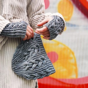 Puede incluir: Un gorro de punto gris con un patrón de trenza. El gorro está siendo sostenido por una persona que lleva un suéter blanco y guantes sin dedos de punto gris.
