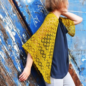 Può includere: Uno scialle di uncinetto giallo con un motivo a pizzo. Lo scialle è drappeggiato sulle spalle di una persona e viene indossato con una blusa blu scuro e pantaloni grigi chiari.