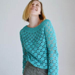 Puede incluir: Un suéter de crochet turquesa de corte holgado y mangas largas. El suéter tiene un delicado patrón de calado.