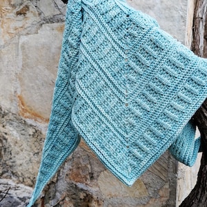 May include: A light blue crochet shawl with a textured, geometric pattern. The shawl is draped over a tree branch, showcasing its intricate stitchwork and soft color. The handmade accessory is a cozy and stylish addition to any wardrobe.