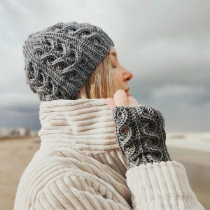 May include: A gray crocheted beanie with a cable knit pattern and matching fingerless gloves. The beanie has a slouchy fit and the gloves have a decorative cable knit pattern.