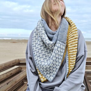 Puede incluir: Un chal triangular de ganchillo hecho a mano en tonos de azul, blanco y amarillo. El chal presenta un patrón de puntadas texturizadas y se lleva alrededor del cuello y los hombros. El fondo muestra una playa y un cielo nublado.