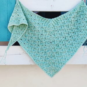 Può includere: Un scialle all'uncinetto color azzurro con un motivo geometrico. Lo scialle è drappeggiato su una superficie di legno bianco.