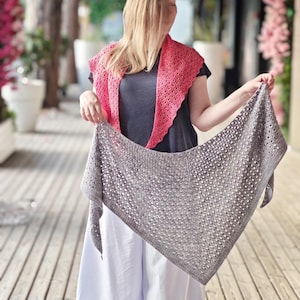 May include: A triangular grey knitted shawl with an openwork lace pattern held in front of a dark grey t-shirt and a bright pink knitted shawl draped over the shoulders. The grey shawl features a textured stitch design with visible yarn details.