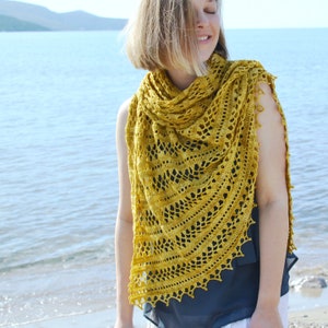 May include: A woman wearing a yellow crochet shawl with a lacy pattern. The shawl is draped over her shoulders and is tied at her neck. She is standing on a beach with the ocean in the background.