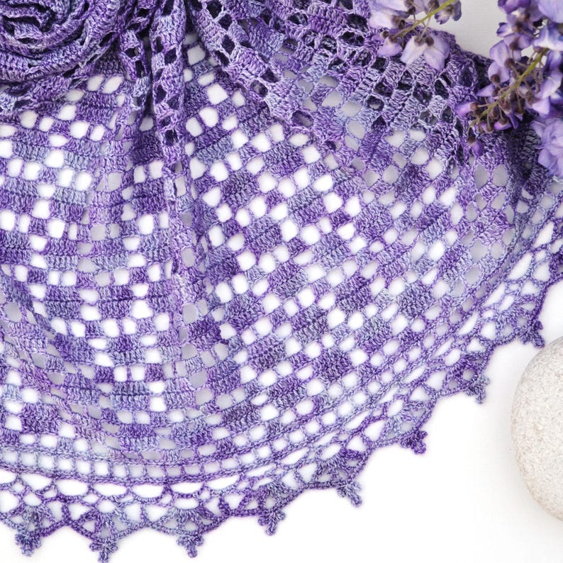 Puede incluir: Un chal de crochet hecho a mano, de tejido abierto, en tonos morados. El chal presenta un patr&oacute;n geom&eacute;trico con un borde festoneado. La tela suave y ligera est&aacute; drapeada, mostrando el intrincado dise&ntilde;o. Una ramita de flores moradas est&aacute; en el fondo.