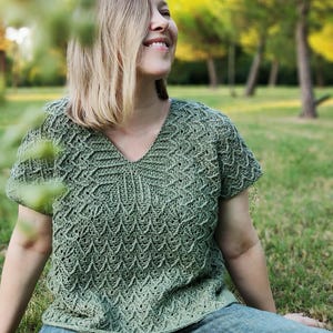 Könnte beinhalten: Ein grünes Häkeltop mit V-Ausschnitt. Das Oberteil hat kurze Ärmel und ein strukturiertes Muster. Die Person trägt eine blaue Jeans und sitzt auf dem Gras.
