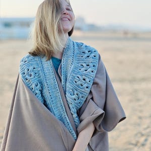 May include: A light blue crocheted shawl with an intricate openwork design is draped over a taupe-colored wrap. The shawl is worn over a teal shirt. The image is taken outdoors, with a blurred background of a beach and sky.