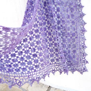 Puede incluir: Un chal hecho a mano, de tejido abierto, en tonos morados y lavanda. El chal presenta un patr&oacute;n de cuadros y un borde festoneado. El dise&ntilde;o delicado sugiere una sensaci&oacute;n ligera y aireada, perfecta para superponer.