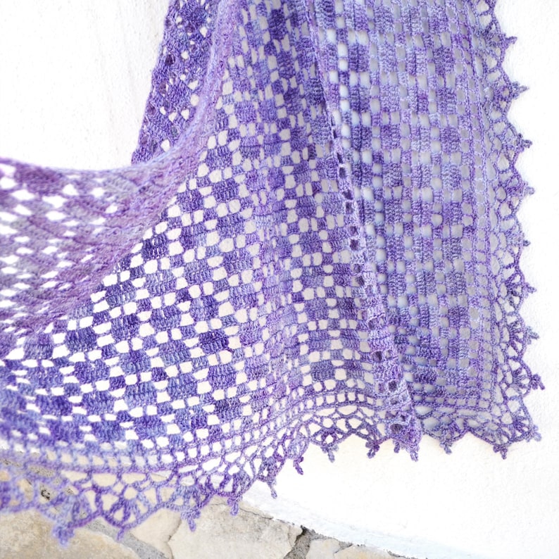 Puede incluir: Un chal hecho a mano, de tejido abierto, en tonos morados y lavanda. El chal presenta un patr&oacute;n de cuadros y un borde festoneado. El dise&ntilde;o delicado sugiere una sensaci&oacute;n ligera y aireada, perfecta para superponer.