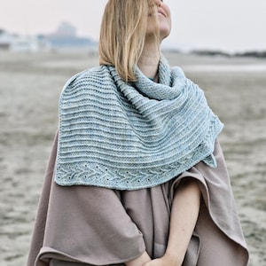 May include: A light blue knitted shawl with a textured pattern and a decorative border is draped over a beige cape. The shawl is wrapped around the neck and shoulders. The background is a beach scene.