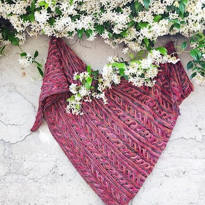 Peut inclure: Un châle tricoté de couleur rouge, rose et violet, avec un motif en tricot torsadé. Le châle est drapé contre un mur texturé et orné de fleurs blanches et de feuilles vertes.