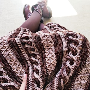 Könnte beinhalten: Eine gehäkelte Decke in Braun und Rosa mit einem Zopfmuster. Die Decke ist gefaltet und über die Beine einer Person gelegt.