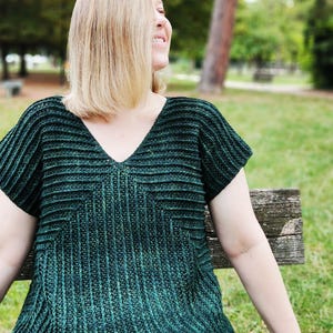 May include: A dark green, short-sleeved top with a V-neck. The top has a ribbed texture and a geometric design on the front. The person is sitting on a wooden bench in a park.