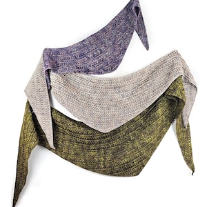 May include: Three knitted shawls in shades of purple, tan, and olive green hang against a white background. Each shawl features a textured, chevron-like pattern. The shawls are draped to showcase their shape and design.