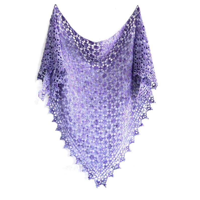 Puede incluir: Un chal hecho a mano, de tejido abierto, en tonos morado, lavanda y blanco. El chal triangular presenta un delicado patr&oacute;n de encaje y un borde festoneado. El chal se muestra sobre un fondo blanco, destacando su intrincado dise&ntilde;o.