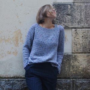 Pode incluir: Uma mulher vestindo um suéter azul claro com um brilho sutil e calças de riscas azuis escuras. Ela está em pé contra uma parede com textura de pedra.