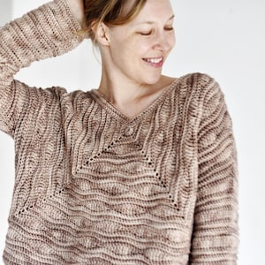 Puede incluir: Un suéter de punto beige y marrón con cuello en V. El suéter presenta un patrón texturizado en forma de onda y mangas largas. La prenda de punto es de color neutro, adecuada para varios conjuntos.