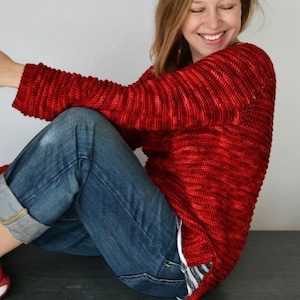 May include: A woman wearing a red crocheted sweater and blue jeans. The sweater has a loose fit and a slightly slouchy neckline.