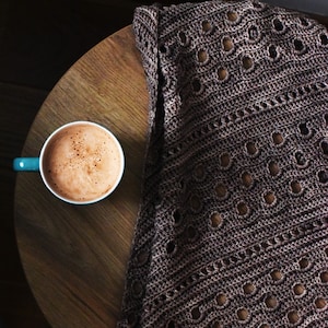 May include: A cup of hot chocolate with a brown crocheted blanket on a wooden table.