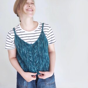 May include: A teal blue crocheted tank top with a cable knit pattern. The tank top has thin straps and a v-neck.