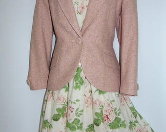 Puede incluir: Una americana rosa empolvado con cierre de un botón y dos bolsillos con solapa, sobre un vestido color crema con estampado floral verde y rosa, falda de volantes y delicado detalle de encaje en el dobladillo.