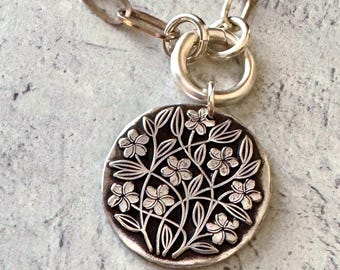 May include: A silver-toned necklace featuring a circular pendant with an embossed floral pattern of small flowers and leaves. The pendant is attached to a thick, oval-linked chain by a large, smooth circular jump ring connector.