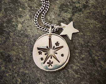May include: A silver necklace with a round pendant featuring a starburst design with smaller stars around it. A smaller silver star charm hangs from the pendant.